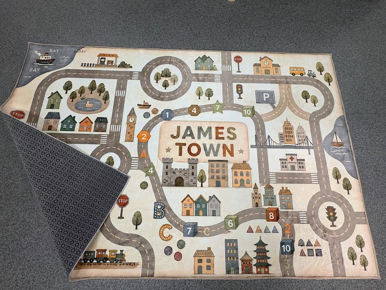 May include: A rectangular play rug featuring a printed town map with roads, buildings, trees, and a train. The center displays the text JAMES TOWN. The rug has a light-colored top surface with a detailed city graphic and a dark, patterned underside.