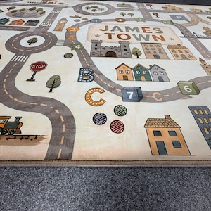 May include: A rectangular area rug featuring a printed town map labeled JAMES TOWN. The design includes winding gray roads, a train, various houses, buildings, trees, a stop sign, and educational elements like letters and numbered blocks.