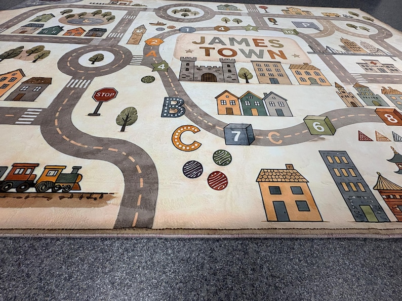May include: A rectangular area rug featuring a printed town map labeled JAMES TOWN. The design includes winding gray roads, a train, various houses, buildings, trees, a stop sign, and educational elements like letters and numbered blocks.