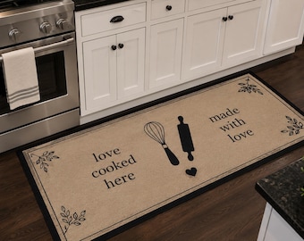 Può includere: Tappeto da cucina beige con bordo nero, con frusta, mattarello e grafica a cuore. Il testo recita "love cooked here" e "made with love". Foglie decorative in ogni angolo. Posizionato davanti a un forno in acciaio inossidabile.