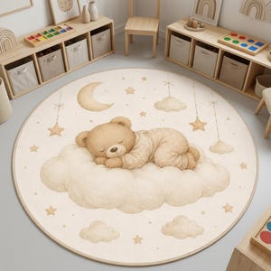 Op de afbeelding: Een rond vloerkleed in een crèmekleur met een centrale illustratie van een bruine teddybeer die op een grote wolk slaapt. Het ontwerp bevat een halve maan, verschillende kleine sterren en extra wolken die aan dunne lijnen hangen.