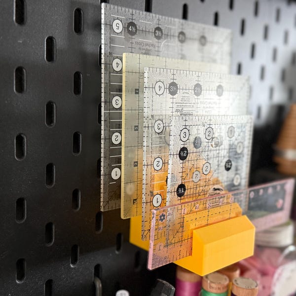May include: A collection of clear acrylic quilting rulers with black markings, stacked and held by a bright yellow holder. The rulers are marked with numbers and angles for precise measurements. The rulers are mounted on a black pegboard.