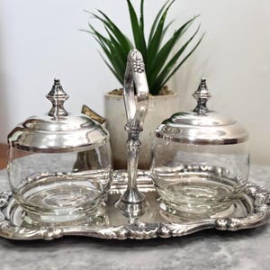 Peut inclure: Un service à condiments argenté composé de deux bols en verre transparent avec des couvercles en argent assortis. L'ensemble est disposé sur un plateau ovale en argent doté d'une poignée centrale verticale avec un motif en relief.