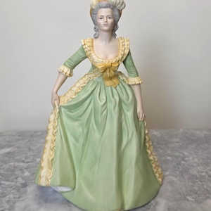 Puede incluir: Una figura de porcelana de una persona con un vestido verde pálido con adornos de volantes amarillos, un lazo amarillo a juego en el corpiño y un peinado gris elaborado y alto adornado con un accesorio amarillo, en una pose formal.