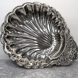 Puede incluir: Una bandeja de servicio de metal plateado pulido con forma de concha de vieira grande. La bandeja presenta surcos profundos y radiantes en toda su superficie y un borde decorativo en relieve con motivos florales y volutas en un lado.