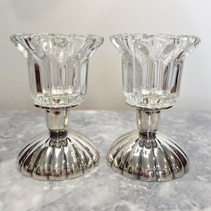 Peut inclure: Une paire de bougeoirs composée de coupelles en verre transparent cannelé reposant sur des bases en métal argenté poli au design festonné en forme de coquille. Chaque bougeoir possède une base large et arrondie et un sommet en verre nervuré.