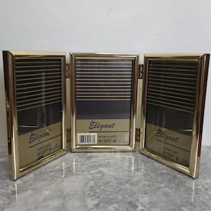 Könnte beinhalten: Ein dreiteiliger Fotorahmen aus goldfarbenem Metall mit drei Scharnierpaneelen. Jeder Rahmeneinsatz zeigt schwarze und goldene horizontale Streifen, das Wort Elegant und den Markennamen Intercraft, mit sichtbarem 10x15cm Etikett.