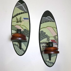 Peut inclure: Deux appliques murales ovales présentant des paysages peints à la main avec des collines vertes, des montagnes grises et des petites maisons. Chaque applique possède une étagère à bougie en bois brun foncé avec une coupelle noire au centre.