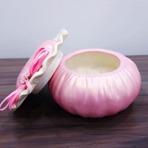 May include: A pink, round trinket box with a matching lid. The box has a ribbed texture and a white rim on the lid. Pink ribbons and a small flower adorn the lid. The box is open, revealing a light interior.