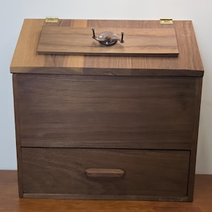 May include: A dark brown wooden storage box with a slanted top lid featuring a metal handle and two brass hinges. The front of the box includes a single horizontal drawer with a matching wooden handle, showcasing a smooth, natural wood grain finish.