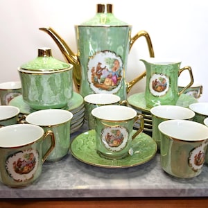 May include: A light green porcelain tea set featuring gold trim and ornate central illustrations of figures in period clothing. The set includes a tall teapot, a lidded sugar bowl, a creamer, and multiple matching cups and saucers.