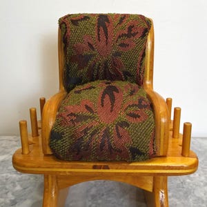May include: A miniature wooden chair-shaped spool holder with a polished light brown finish. The seat and backrest are upholstered in a textured fabric featuring a dark green and brown floral pattern. Six vertical wooden pegs surround the base.