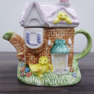 May include: A decorative ceramic teapot in the shape of a house. It features a pink roof, brickwork walls, and a green handle. A yellow chick is perched on the grass. The teapot has a whimsical design.