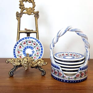 May include: A decorative brass easel holding a small round ceramic plate with blue and pink floral patterns, alongside a matching ceramic basket holder containing a stack of five small round ceramic plates with identical blue and pink floral designs.
