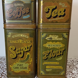 May include: Four rectangular metal storage canisters with lids, featuring vintage-style labels for Coffee, Tea, Sugar, and Flour. The containers are coloured in shades of olive green, brown, and gold, with decorative borders and text printed on each.