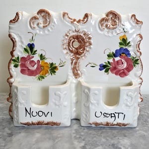 May include: A white ceramic wall-mounted double pocket organiser with brown scrollwork edges. The front features hand-painted floral motifs in pink, yellow, blue, and green. The left pocket is labelled Nuovi and the right pocket is labelled Usati.