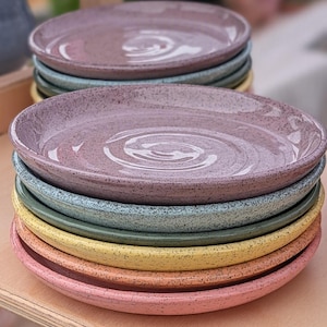 May include: A stack of round ceramic plates with a speckled finish. The plates feature a variety of colors including mauve, sage green, yellow, orange, and pink. Each plate has a slightly raised rim and a smooth, glossy surface texture.