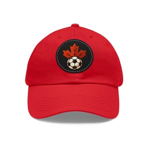 May include: A red baseball cap with a circular patch showing a soccer ball and a maple leaf. The patch has a black background and a white stitched border. The maple leaf is orange and the soccer ball is black and white.