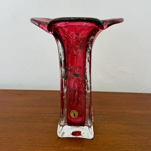 Puede incluir: Un jarrón de cristal rojo rubí con la parte superior acampanada y una base rectangular. El jarrón tiene un aspecto texturizado con detalles oscuros y una pequeña pegatina redonda cerca de la base. El jarrón se exhibe sobre una superficie de madera.