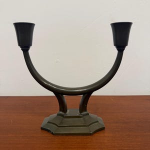 May include: A bronze-coloured, two-candle candelabra. The base is octagonal, and the arms curve upwards to hold the candle cups. The candelabra is made of metal and sits on a wooden surface.