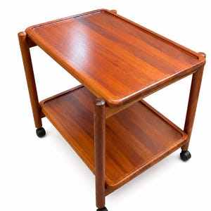 May include: A two-tiered wooden serving trolley with a warm brown finish. The trolley features rectangular shelves with raised edges, supported by four cylindrical corner posts. It is mounted on four black swivel castors for mobility.