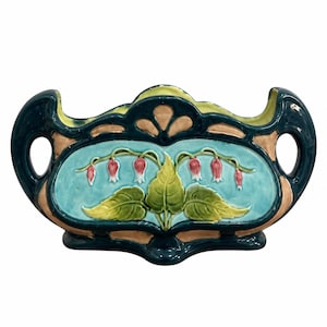 May include: A ceramic planter featuring a dark teal frame with two side handles and a central light blue panel. The panel displays a relief design of green leaves and pink and white bell-shaped flowers. The interior is a pale yellow color.