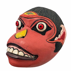 Può includere: Una maschera in legno intagliato a mano con un volto rosso, capelli neri, denti bianchi prominenti e grandi occhi con iridi rosse. La maschera presenta un dettaglio del naso giallo, contorni neri e un piccolo emblema decorativo.