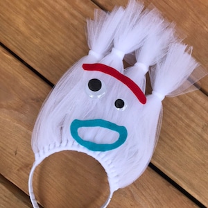 May include: White tulle headband with a felt face, featuring googly eyes, a red mouth, and a blue smile.