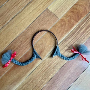 May include: A grey braided headband with two grey tulle bows with red ribbons attached. The headband is on a wooden surface.