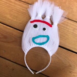 May include: White tulle headband with a felt face, featuring red eyebrows, blue eyes, and a turquoise mouth. The headband is perfect for a costume or party.