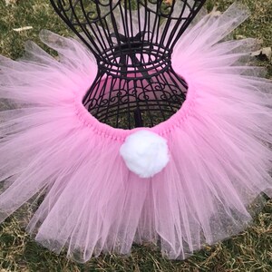 Könnte beinhalten: Ein rosa Tüll-Tutu mit einem weißen Bommel, der an der Vorderseite befestigt ist. Das Tutu befindet sich auf einem schwarzen Mannequin.