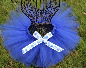 Peut inclure: Un tutu en tulle bleu royal avec un ruban blanc noué en un nœud. Le ruban a un motif répété de toupies bleues et d'étoiles de David.