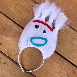 May include: White tulle headband with a felt face, featuring black eyes, a red mouth, and a turquoise smile.