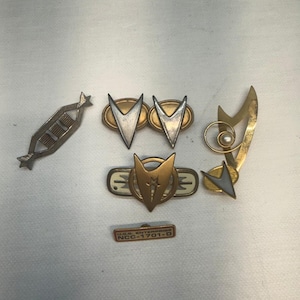 Op de afbeelding: Een verzameling van vijf metalen spelden, waaronder een zilverkleurige geometrische balk, twee ovale spelden met zilveren delta-insignes, een goudkleurig rond embleem met een ster, een abstracte gouden broche met een parel en een rechthoekige U.S.S. ENTERPRISE NCC-1701-D speld.