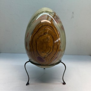 May include: A polished, egg-shaped stone sculpture featuring concentric bands of brown, tan, and translucent green mineral layers. The smooth, rounded object is displayed resting securely within a thin, dark metal wire stand with three curved legs.