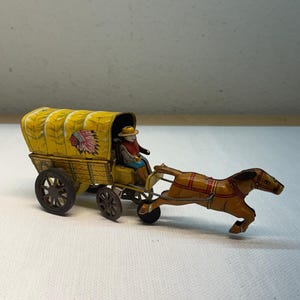 Pode incluir: Um brinquedo de lata em estilo vintage com uma carroça amarela puxada por um cavalo castanho. A cobertura amarela tem um gráfico vermelho e preto de um cocar. Uma pequena figura com chapéu e colete vermelho está sentada na carroça.