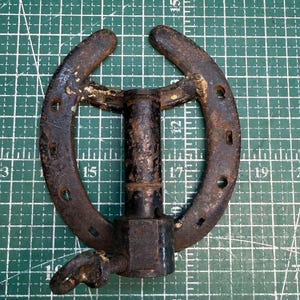 May include: A weathered, dark brown metal horseshoe welded to a central cylindrical post with a square base and a small protruding wing nut. The object shows signs of oxidation and rust, featuring multiple nail holes along the curved horseshoe frame.