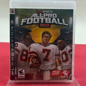 May include: A PlayStation 3 video game case for "All-Pro Football 2K8" featuring three football players in white jerseys with orange accents. The game's title is prominently displayed, along with the "2K Sports" logo. The case is displayed on a clear stand.