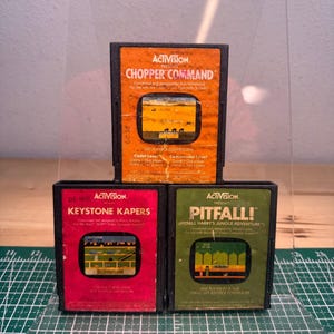Puede incluir: Tres cartuchos de videojuegos vintage Atari 2600 de Activision. El cartucho superior es naranja y está etiquetado como Chopper Command. El inferior izquierdo es rojo y dice Keystone Kapers. El inferior derecho es verde y dice Pitfall!.