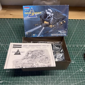 May include: A Monogram SeaQuest DSV Deep Ocean Transport model kit. The box features an illustration of the submersible vehicle. The kit includes parts and instructions. The box is open, showing the contents. Scale model kit.