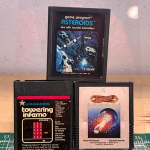 Puede incluir: Tres cartuchos de videojuegos Atari rectangulares de color negro. El cartucho superior es Asteroids. El cartucho inferior izquierdo es Towering Inferno de U.S. Games. El cartucho inferior derecho es Journey Escape. Cada cartucho tiene una etiqueta.