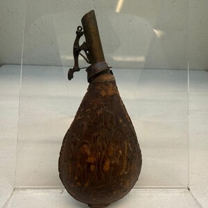 Könnte beinhalten: Eine braune Pulverflasche aus Leder in Tropfenform mit einer Messingtülle. Die Lederoberfläche weist geprägte dekorative Muster auf. Ein federbelasteter Metallmechanismus ist an der Seite des Messingauslaufs zur Pulverabgabe angebracht.