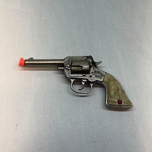 May include: A metallic grey toy revolver featuring a bright orange tip on the barrel. The handle is cream-coloured with an embossed horse head, the text "BUFFALO BILL" in black lettering, and a small circular red accent near the base of the grip.