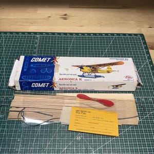 May include: A vintage Comet Aeronca K construction kit. The box features a yellow and orange airplane illustration. Included are wooden strips, a red propeller, and an order form. The kit is designed for building a model airplane.