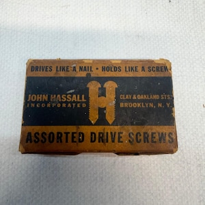 May include: A rectangular cardboard box for John Hassall Incorporated assorted drive screws. The box is tan and black with the text: DRIVES LIKE A NAIL • HOLDS LIKE A SCREW, JOHN HASSALL INCORPORATED, CLAY & OAKLAND STS. BROOKLYN, N. Y., ASSORTED DRIVE SCREWS.