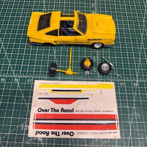 May include: A yellow model car with Monroe and Spraying Systems Co. decals, accompanied by loose axle and wheel components, and a decal sheet featuring the text Over The Road, New York, Detroit, Chicago, Los Angeles, and K-37 on a green cutting mat.