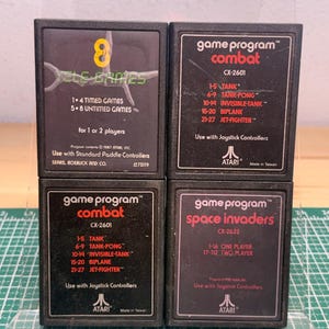 Puede incluir: Cuatro cartuchos de videojuegos Atari rectangulares de color negro. Uno está etiquetado como Tele-Games con 8 juegos, dos como Combat CX-2601 y uno como Space Invaders CX-2632. Cada cartucho presenta texto en blanco y rojo que detalla los modos de juego.