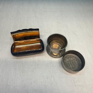 May include: A rectangular black box with a gold-toned interior and a separate silver-toned collapsible cup with a matching lid featuring an embossed shield design. The items are metallic and show signs of age with a smooth, polished finish.