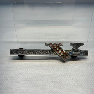 May include: A silver-toned metal emblem featuring the word COMMANDO in raised letters. A large V shape with a red and silver checkered pattern overlaps the bar, and a blue oval with the number 8 is positioned to the right of the V.