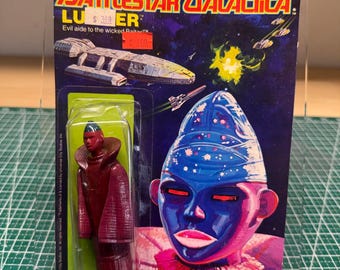 May include: A vintage Battlestar Galactica action figure, still in its original packaging. The packaging features a space scene with a spaceship and the character's name, 'Lurker'. The figure is a red-robed character with a red face and a blue head.
